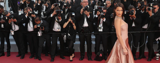 A high-angle shot of bella hadid in a light pink strapless gown walking on a red carpet, surrounded by a line of photographers with cameras raised, capturing her picture.