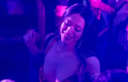 A woman with long dark hair smiles and dances in a dimly lit, purple-and-pink lit club, wearing a low-cut top.